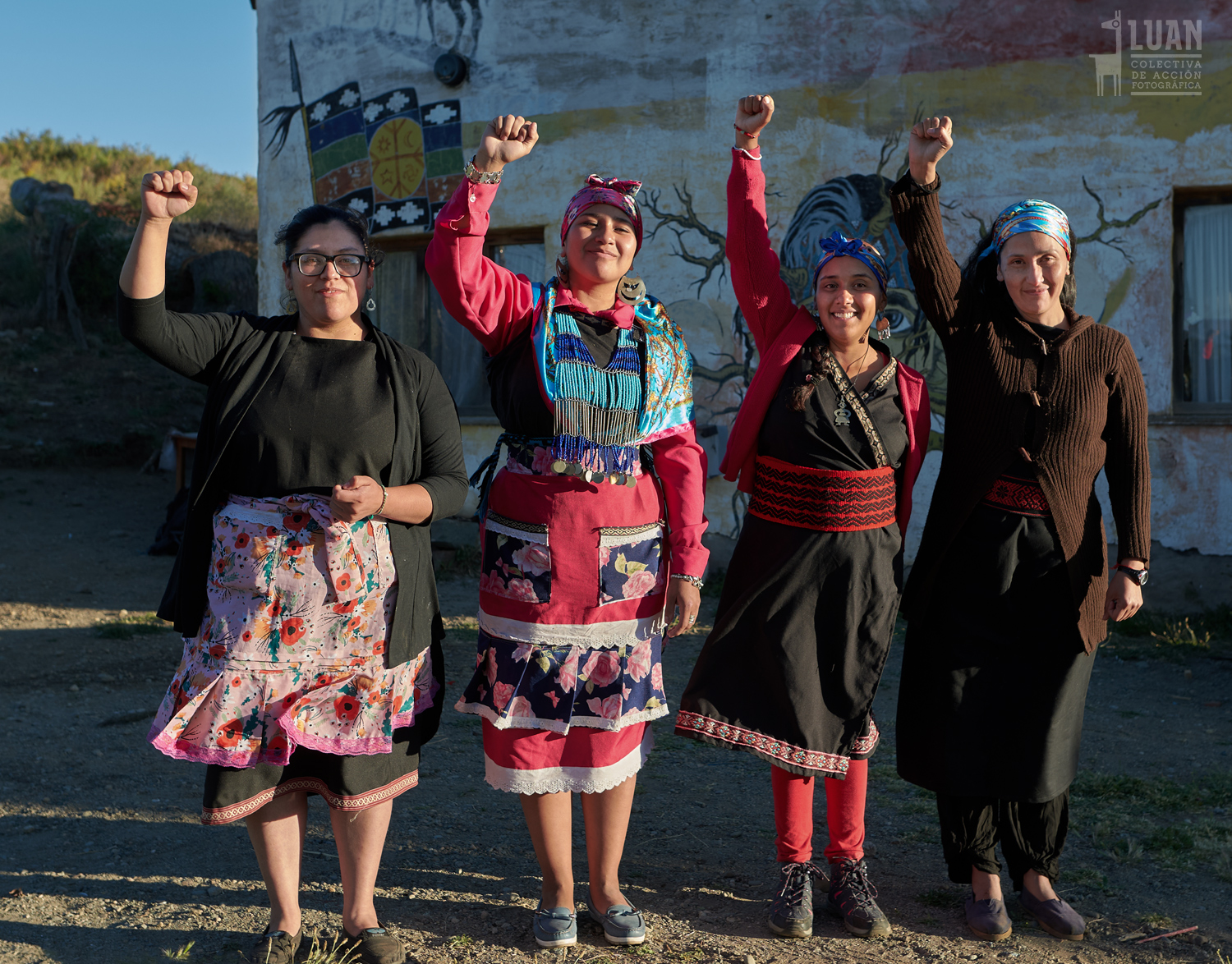 Four women of color stand in a line facing the camera. Each person is smiling and holding up their right fist.